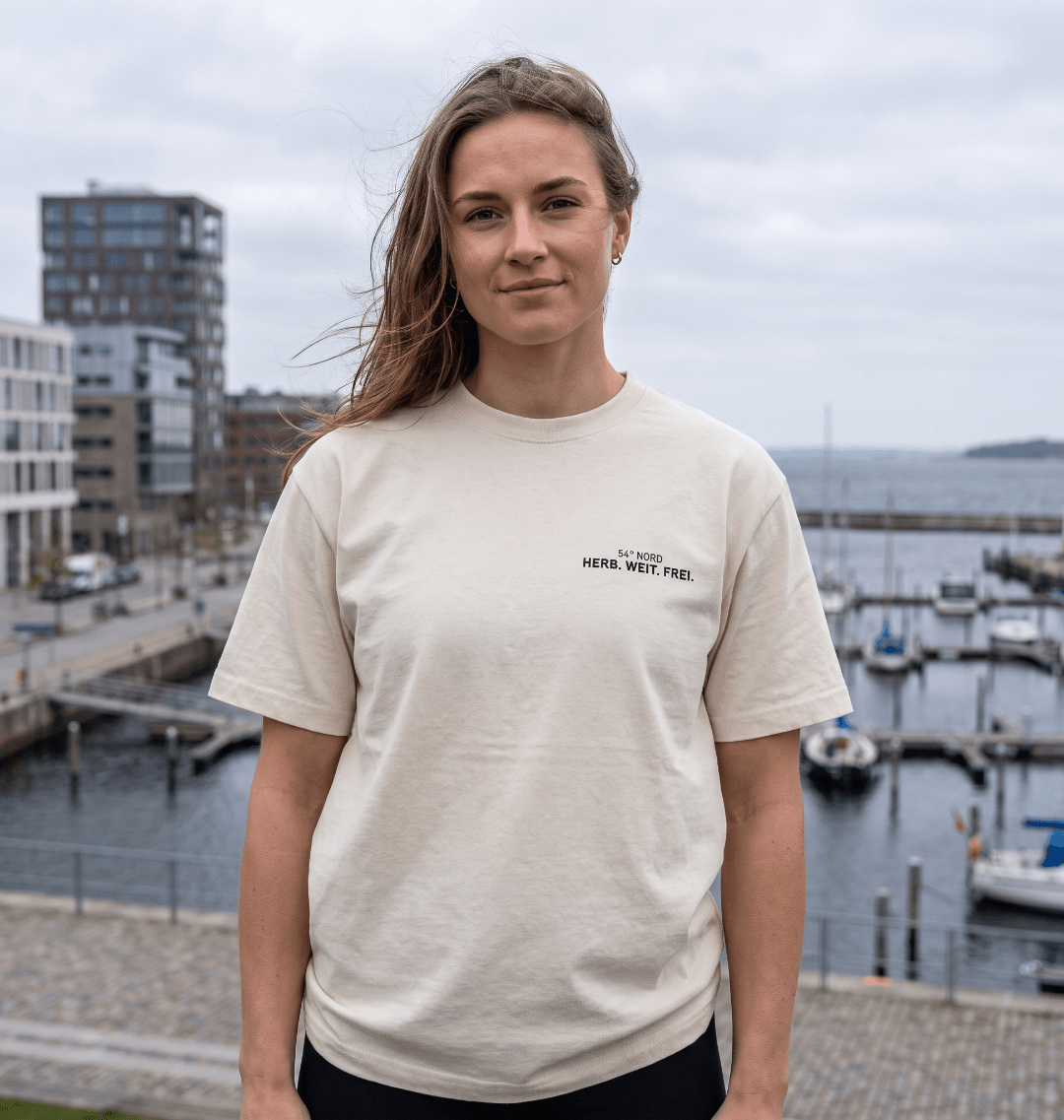 Junge Frau trägt das Herb. Weit. Frei. Oversized T-Shirt in Gischt mit kleinem 54° Nord Brustprint am Wasser.