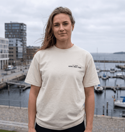Junge Frau trägt das Herb. Weit. Frei. Oversized T-Shirt in Gischt mit kleinem 54° Nord Brustprint am Wasser.