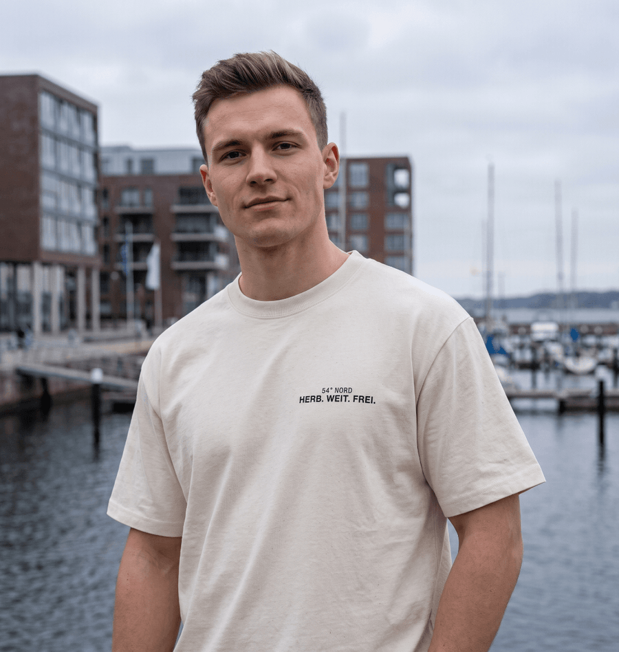 Junger Mann trägt das Herb. Weit. Frei. Oversized T-Shirt in Gischt mit kleinem 54° Nord Brustprint am Hafen.